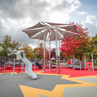 a grey accessible playground with slides and shade on a colourful rubber surface